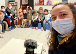 Au premier plan, une adulte avec un micro, et au second plan, un groupe d'enfants de maternelle assis en rond.
