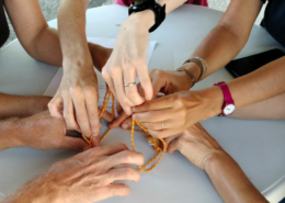 Des mains d'adolescents et d'adultes cherchant ensemble à réaliser une figure avec une cordelette.